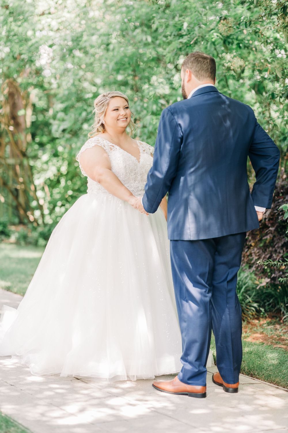 A wedding couple stands together outdoors on a sunny day surrounded by lush green foliage.