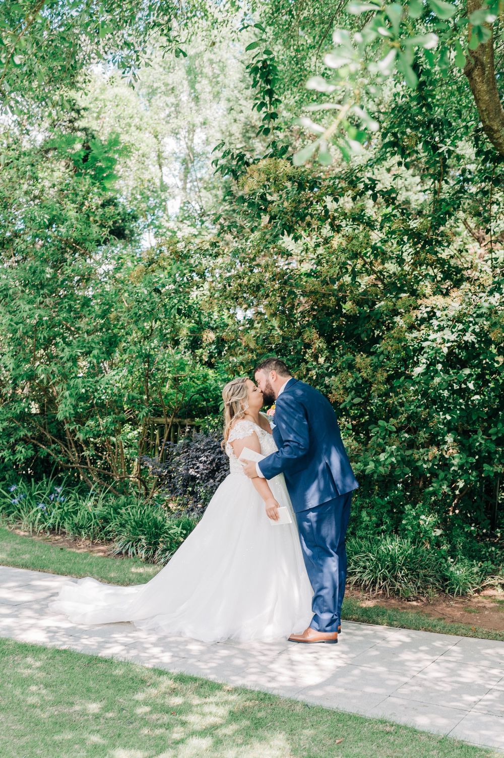 A couple in wedding attire shares an intimate moment outdoors on a sunlit garden path surrounded by lush greenery.