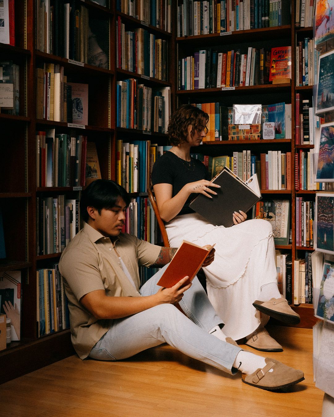 A couple sitting and reading on the floor in a cozy library with floor-to-ceiling bookshelves.
