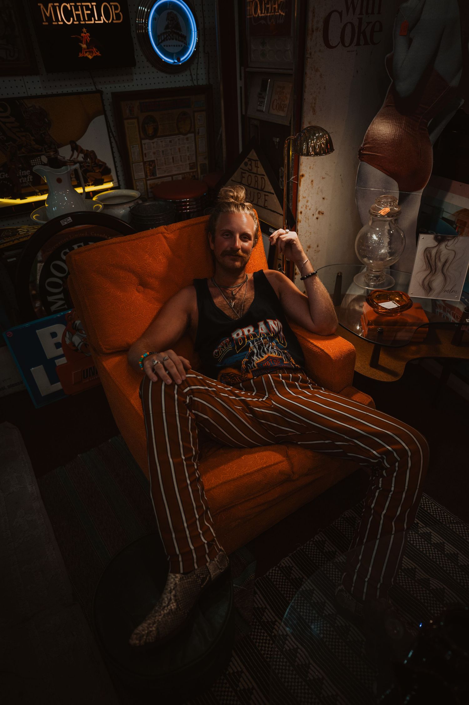 Inside a clothing store with racks of apparel, someone stands posing with a guitar in a dimly lit, moody setting.