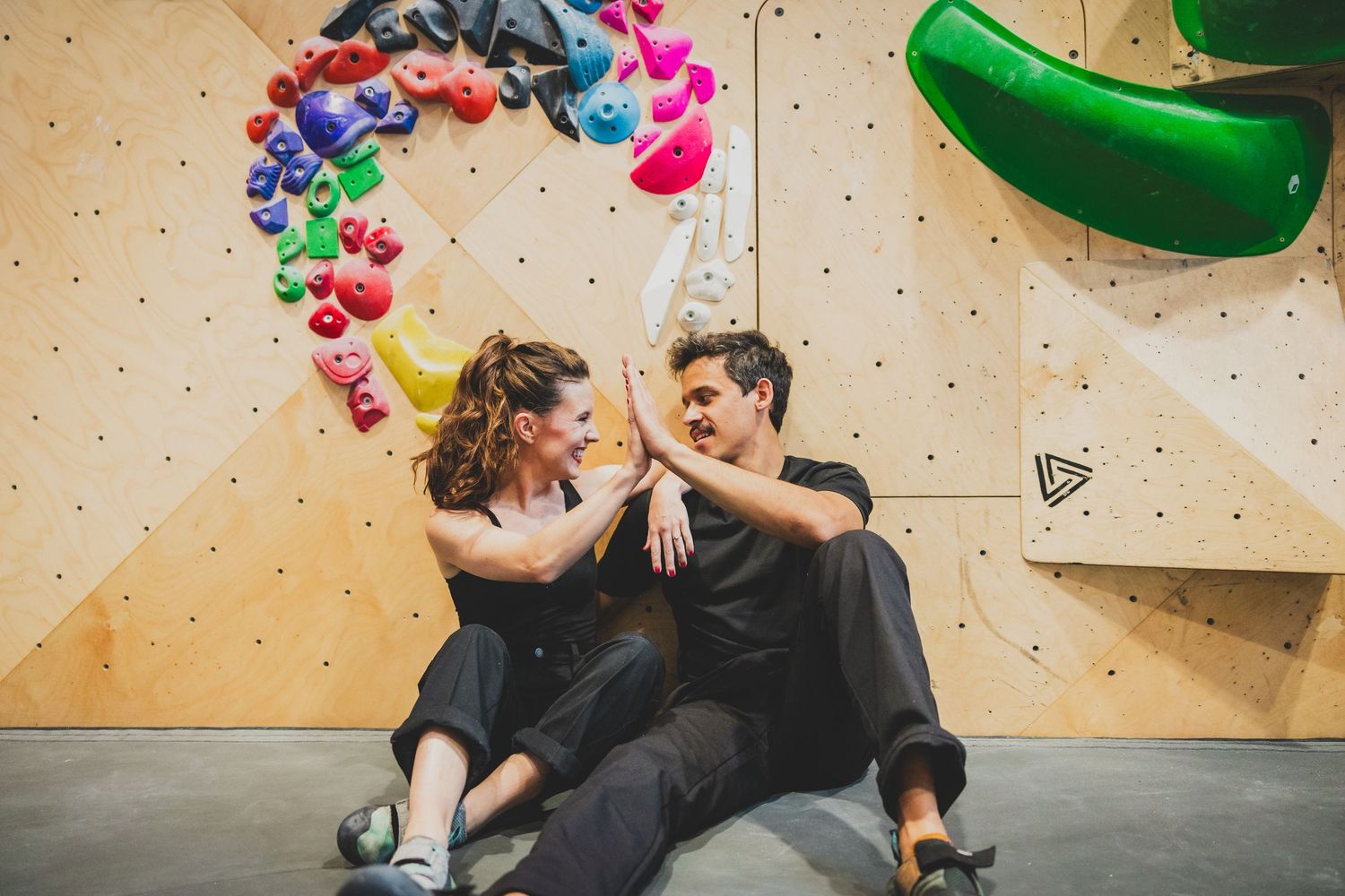 Two people sit together against a climbing wall with colorful heart balloons arranged in an arch above them at the Bouldering Project in DC