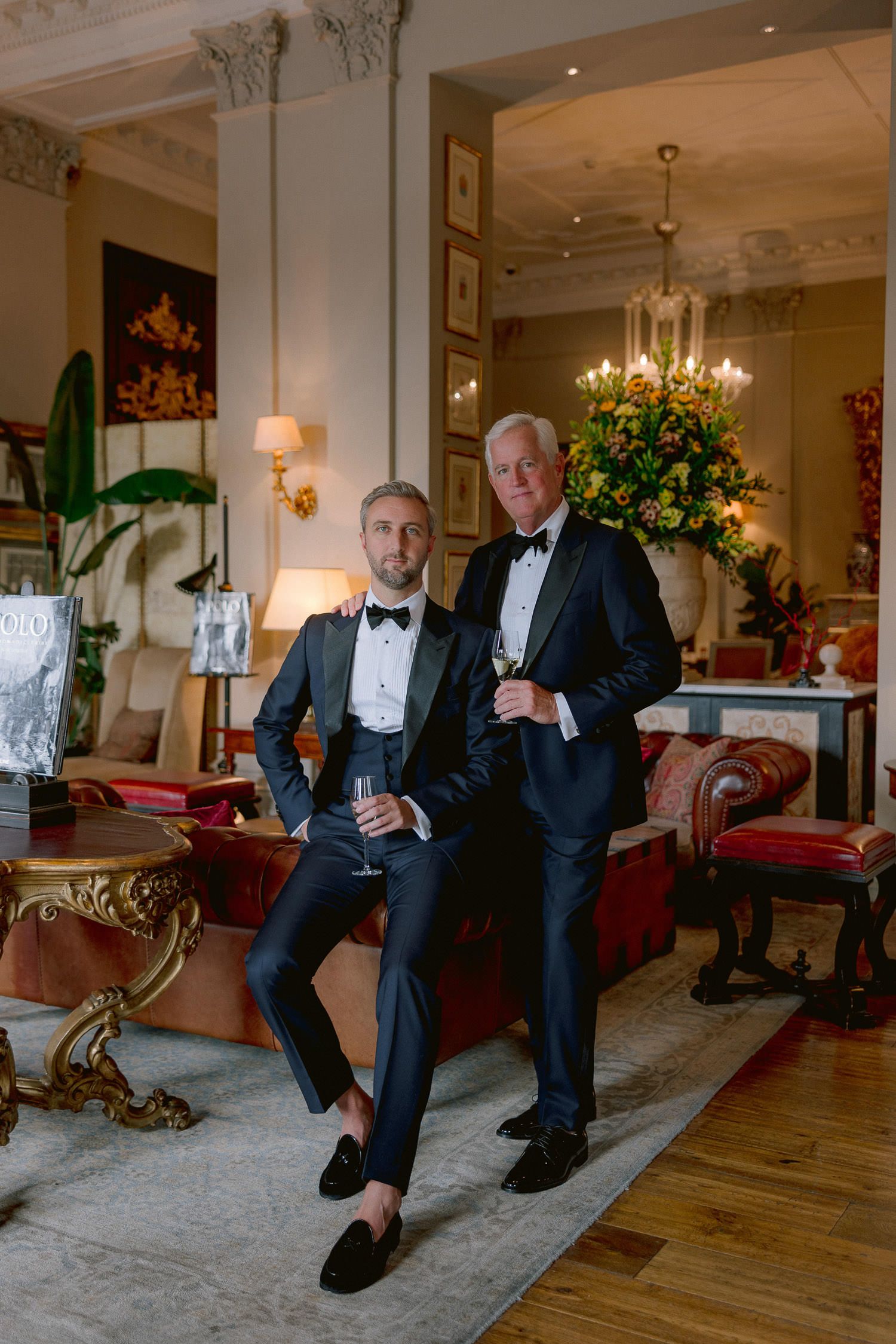 Two people in formal black tuxedos pose in an elegant room with chandelier and ornate furniture.