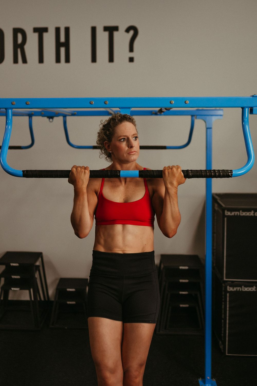 Woman performing pull-ups on a blue exercise bar wearing red sports bra and black shorts.