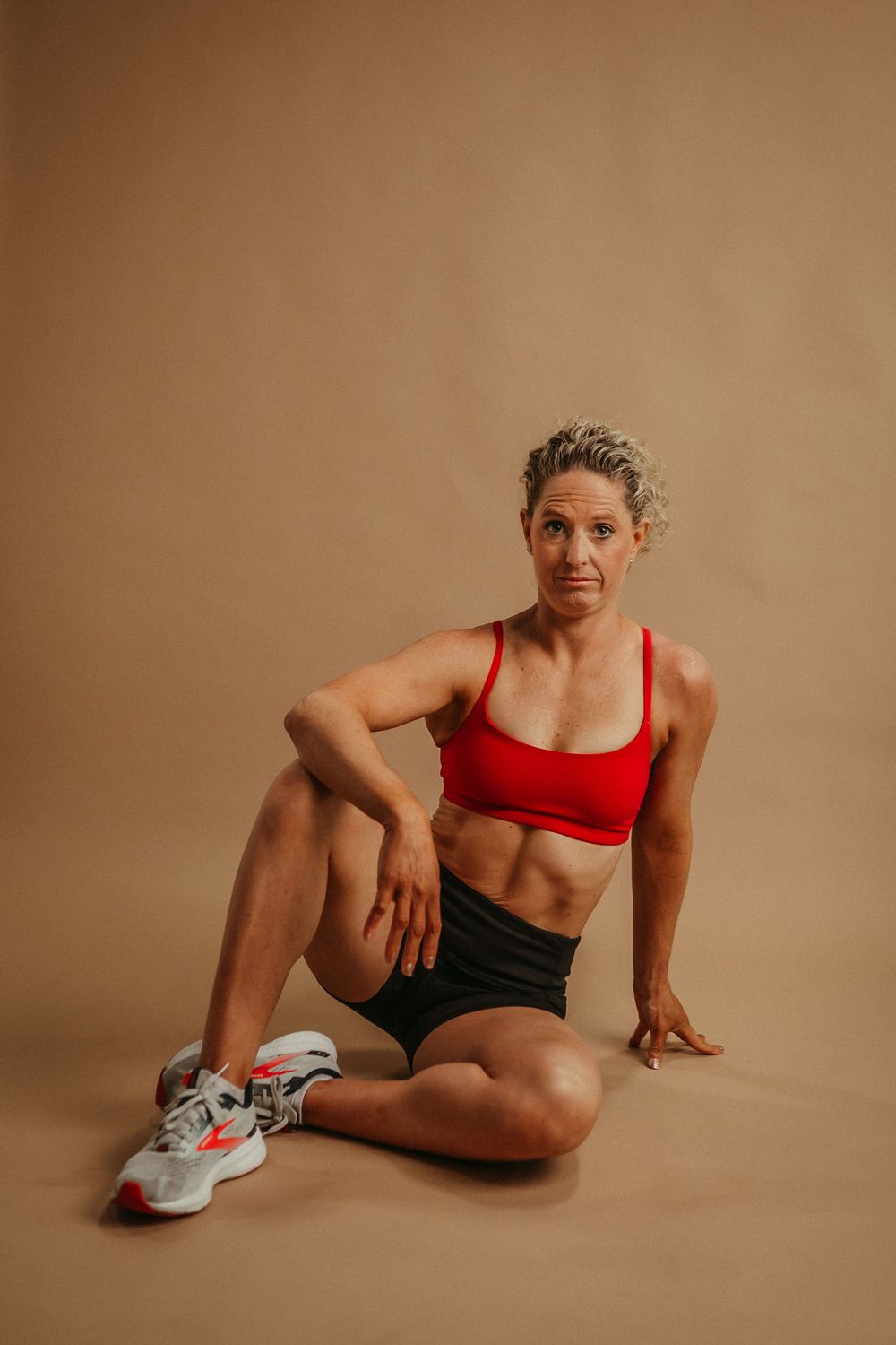 Athletic figure in red sports top and black shorts demonstrating floor stretching poses against beige background.