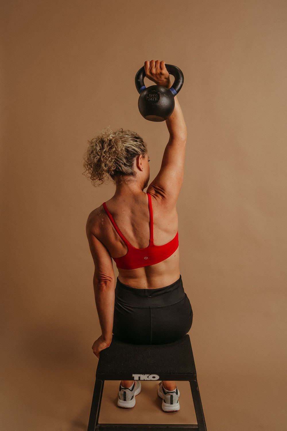 Person in red sports top and black leggings demonstrating proper kettlebell overhead press form in beige studio setting.