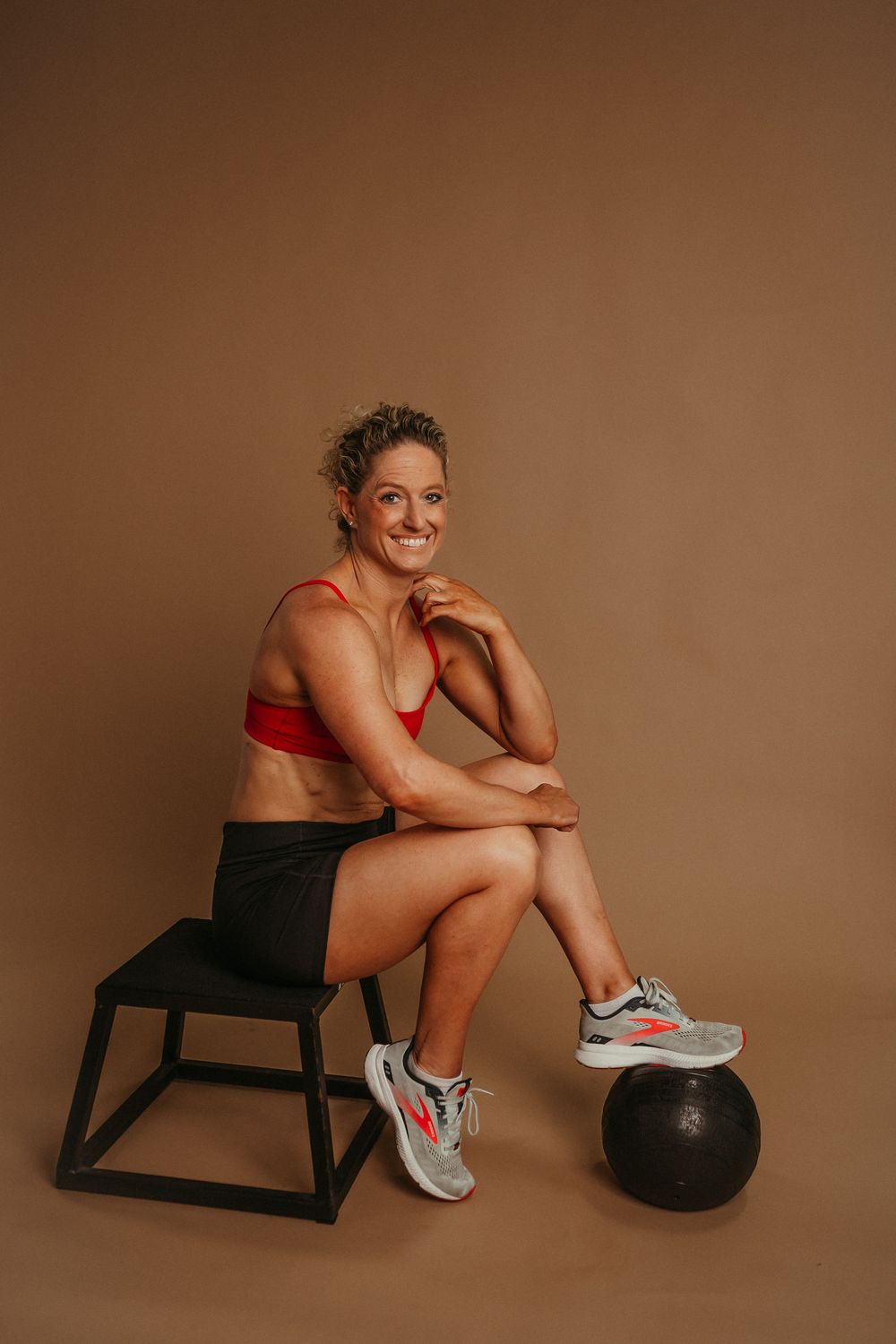 Fitness model rests on black bench with medicine ball against brown backdrop in minimalist studio setting.