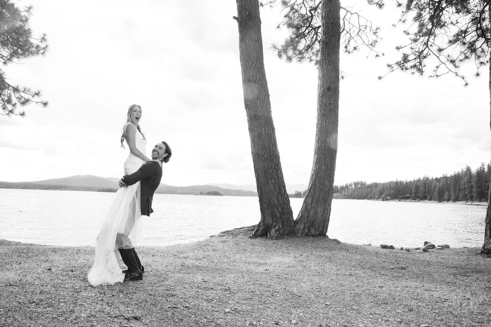 A couple in wedding attire dances and poses in front of a scenic lakeside setting with trees during sunset.
