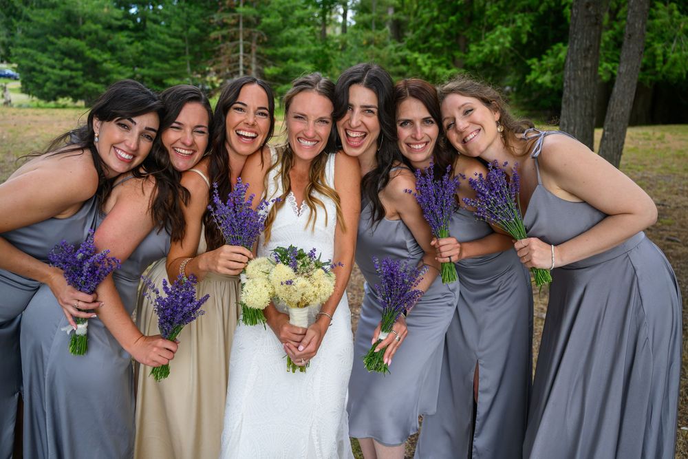 A festive wedding party in gray and beige dresses poses together outdoors holding bouquets of flowers.