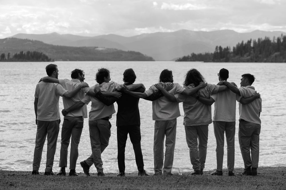 A group of people stand in silhouette with arms around each other while looking out over the water and mountains.