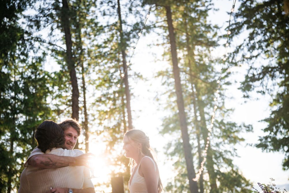 A couple shares an intimate moment in a sunlit forest with tall evergreen trees in the background.