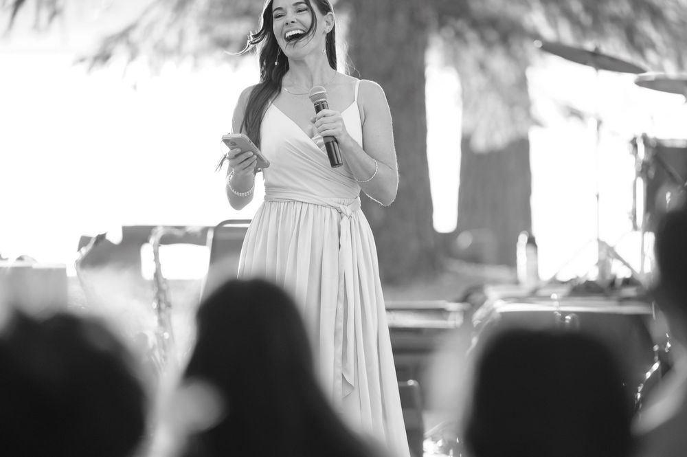 A performer in a light-colored dress stands on stage holding a microphone while performing at an outdoor event.