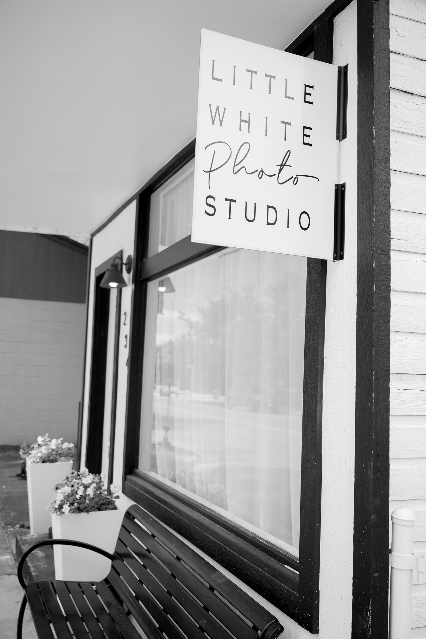 Black and white photograph of a storefront sign reading 'Little White Photo Studio' mounted on brick wall with wooden bench below.