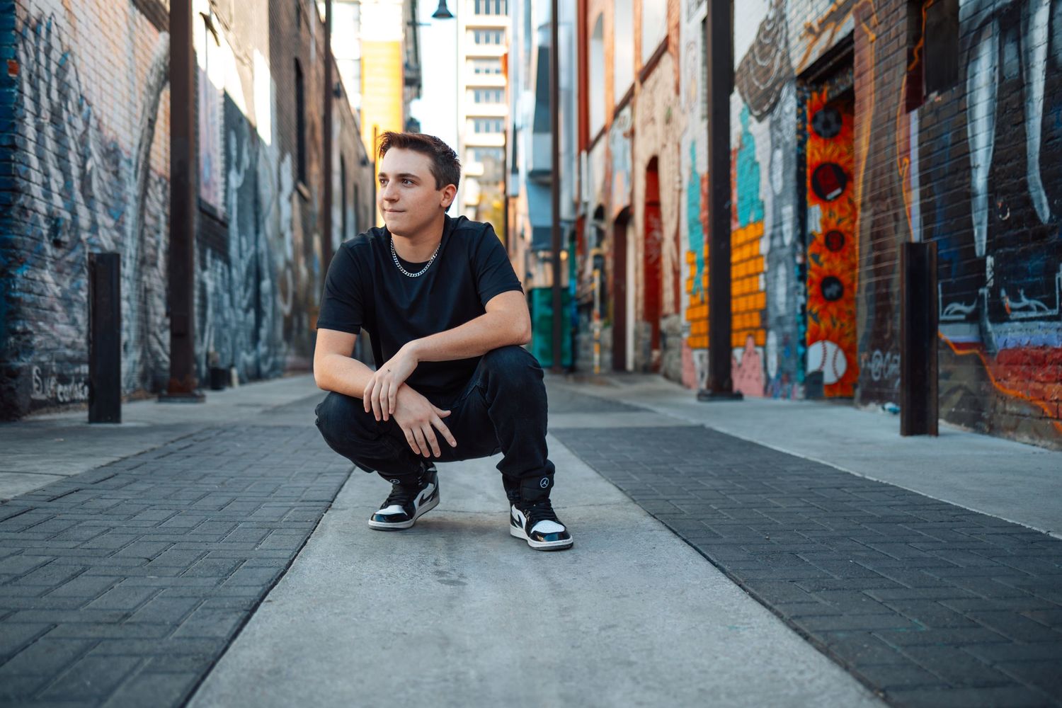 Person in black clothing crouches in urban alleyway with graffiti covered walls and modern architecture in background.