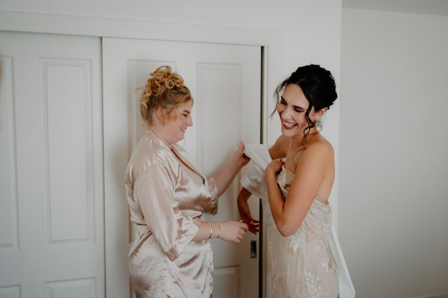 Two women share an intimate bridal preparation moment in a light-filled room wearing satin robes and wedding attire.