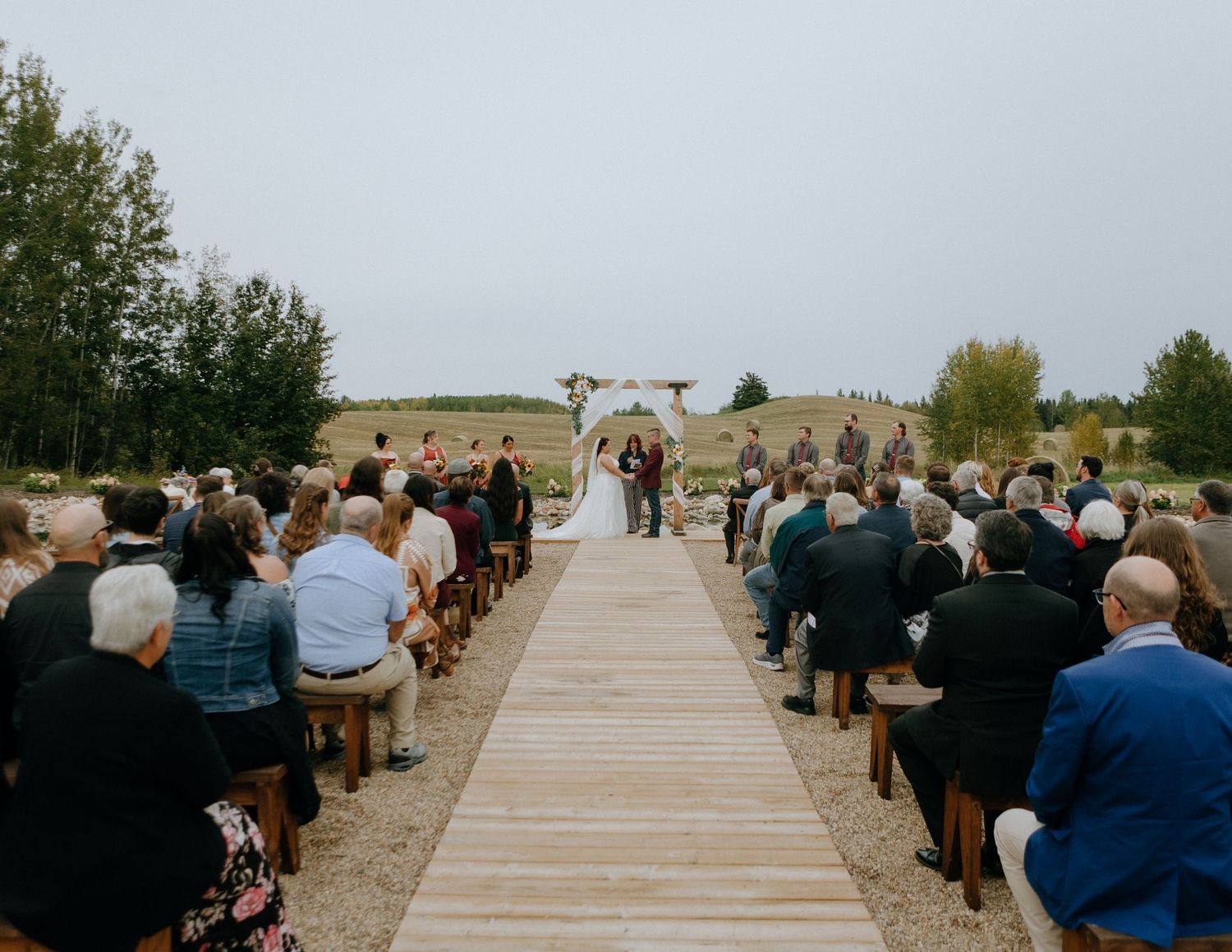 Outdoor wedding ceremony with wooden boardwalk aisle and guests seated on benches.