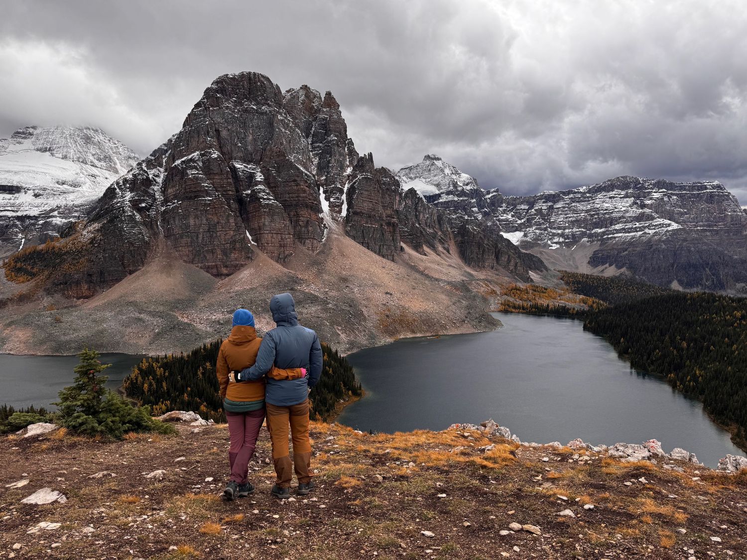 A couple in colorful jackets stand at a viewpoint overlooking Mount Assiniboine and alpine lakes in the Canadian Rockies on an elopement