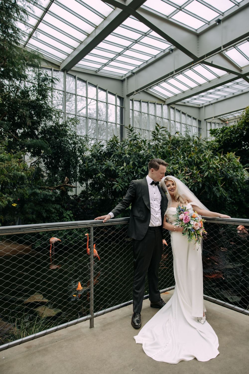 A newlywed couple stands in a glass greenhouse with natural light streaming through the transparent ceiling panels.