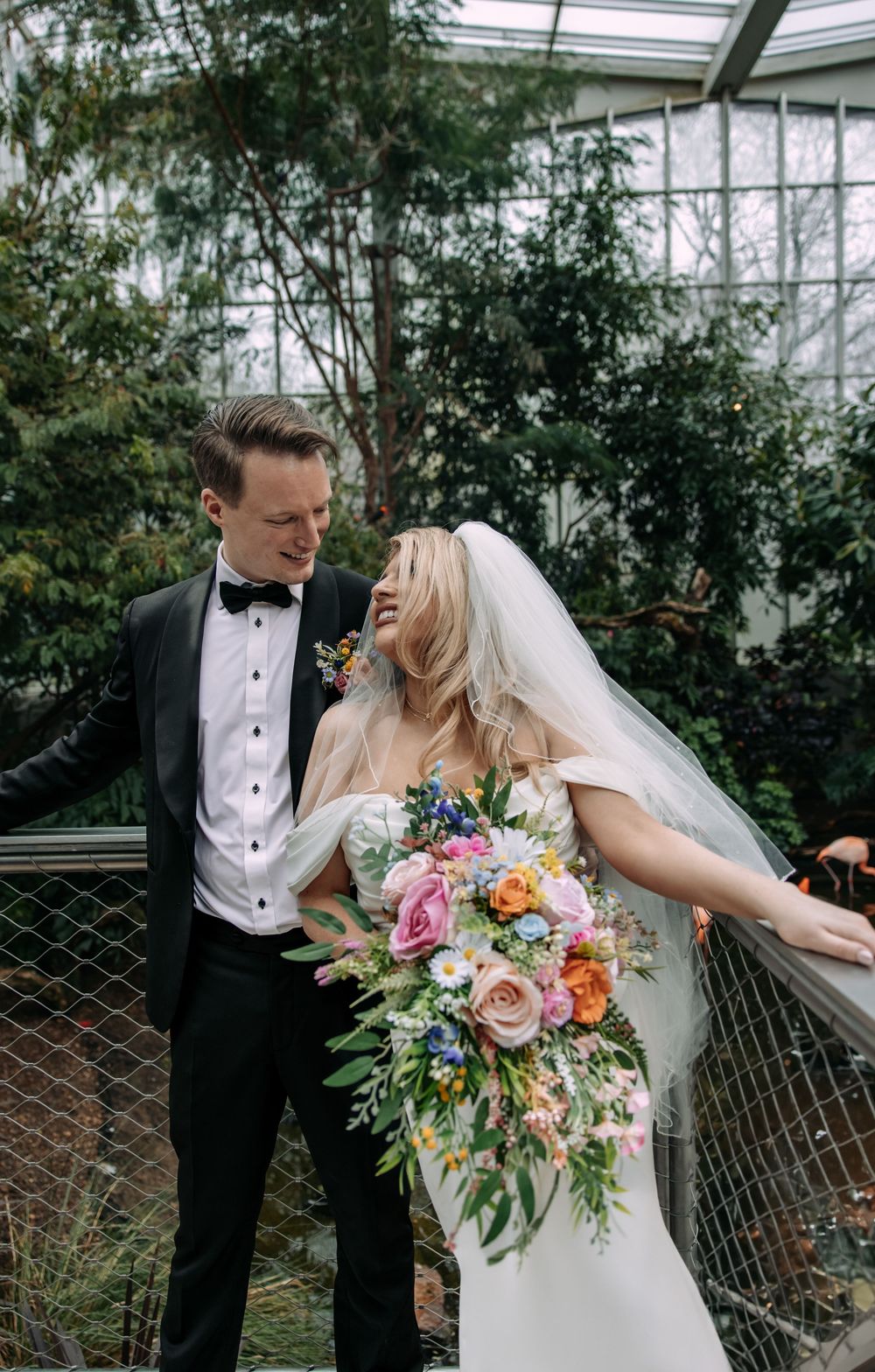 Wedding couple shares joyful moment in greenhouse with lush florals and cascading bridal bouquet.