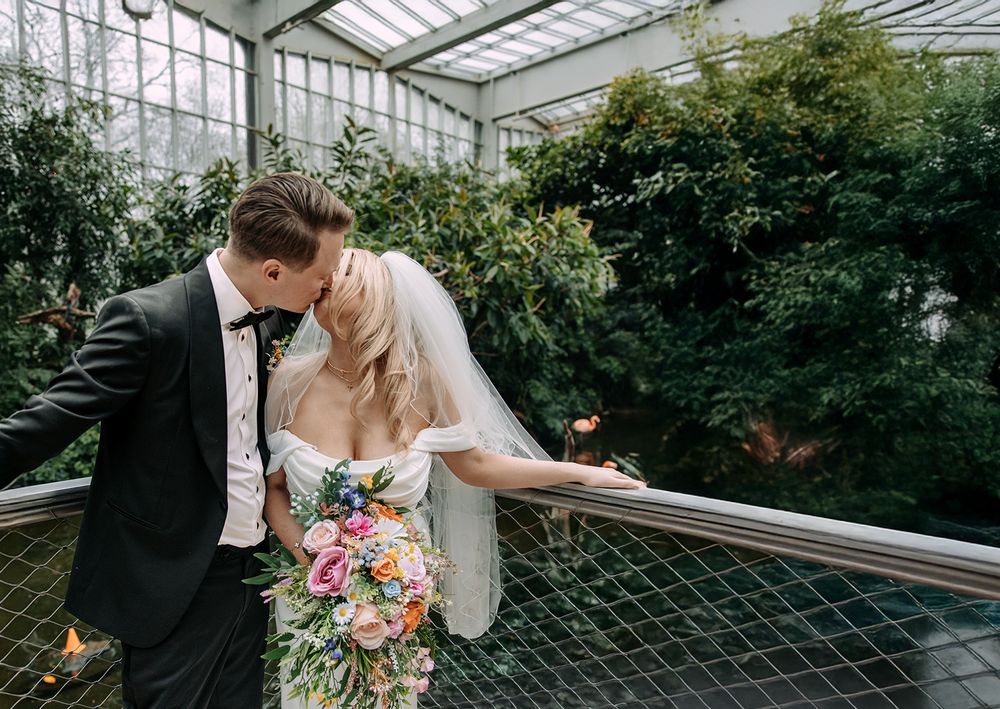 A romantic wedding moment captured in a lush greenhouse conservatory with white veil and colorful bouquet details.