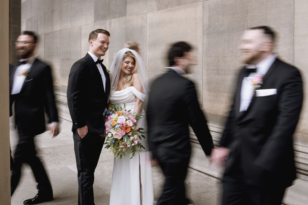 A bride and groom pose against a wall while others in black suits create motion blur walking past them.
