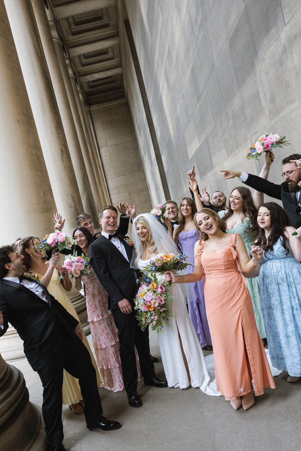 Wedding party celebrates joyfully under grand columned architecture with colorful dresses and floral bouquets.