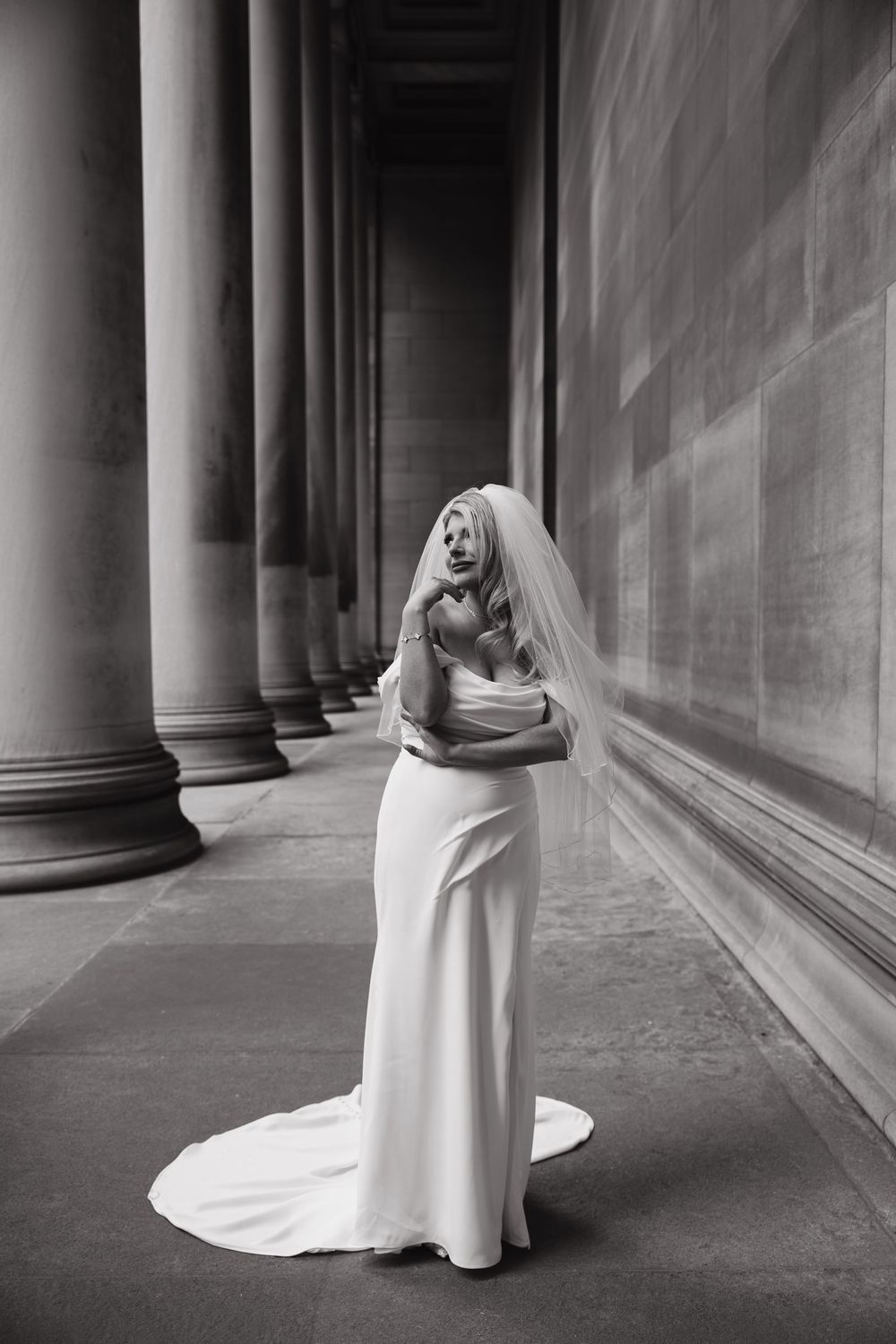 A black and white portrait of a bride in a flowing white dress standing elegantly between classic columns.