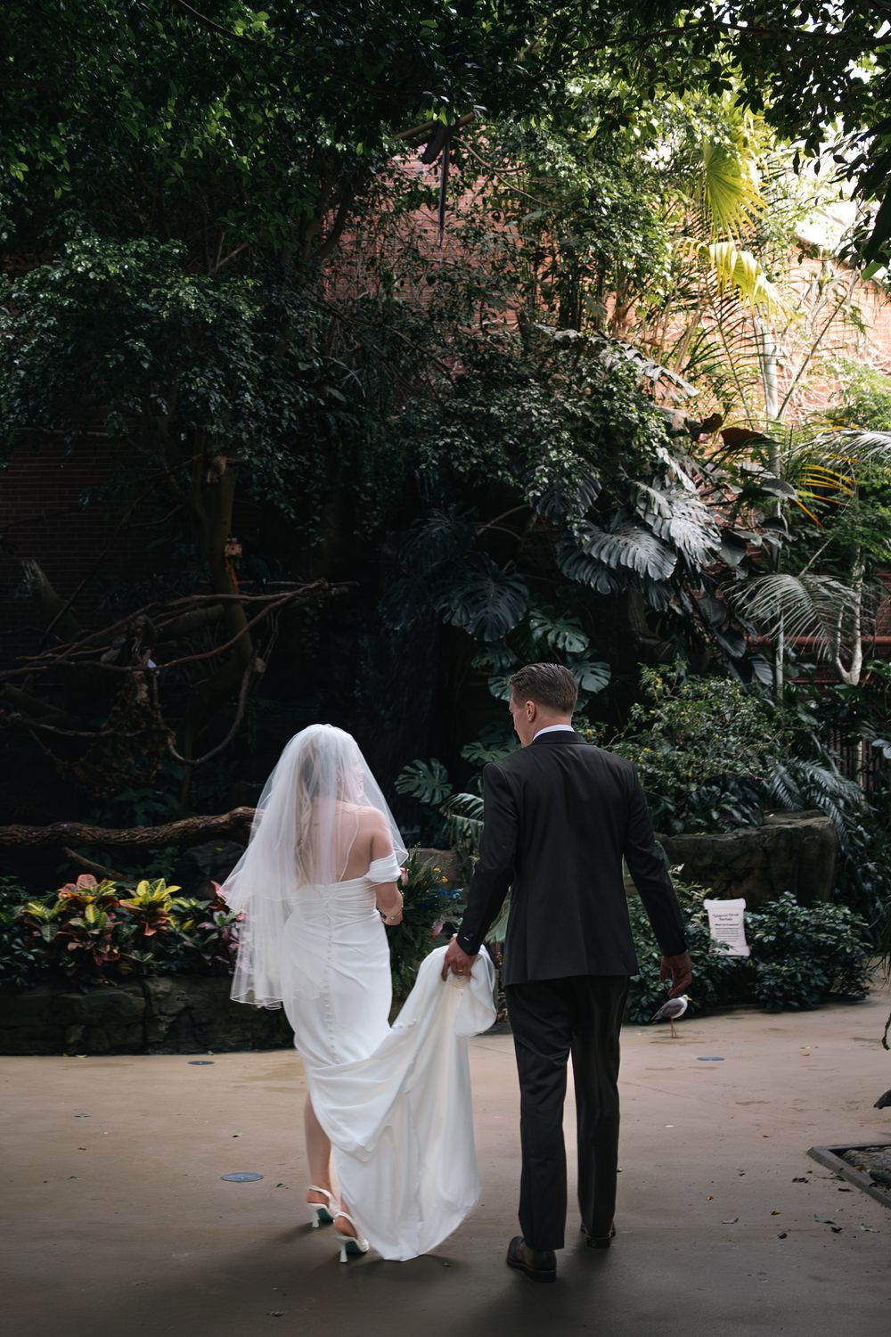 A couple in wedding attire walks along a garden path surrounded by lush greenery and flowers.
