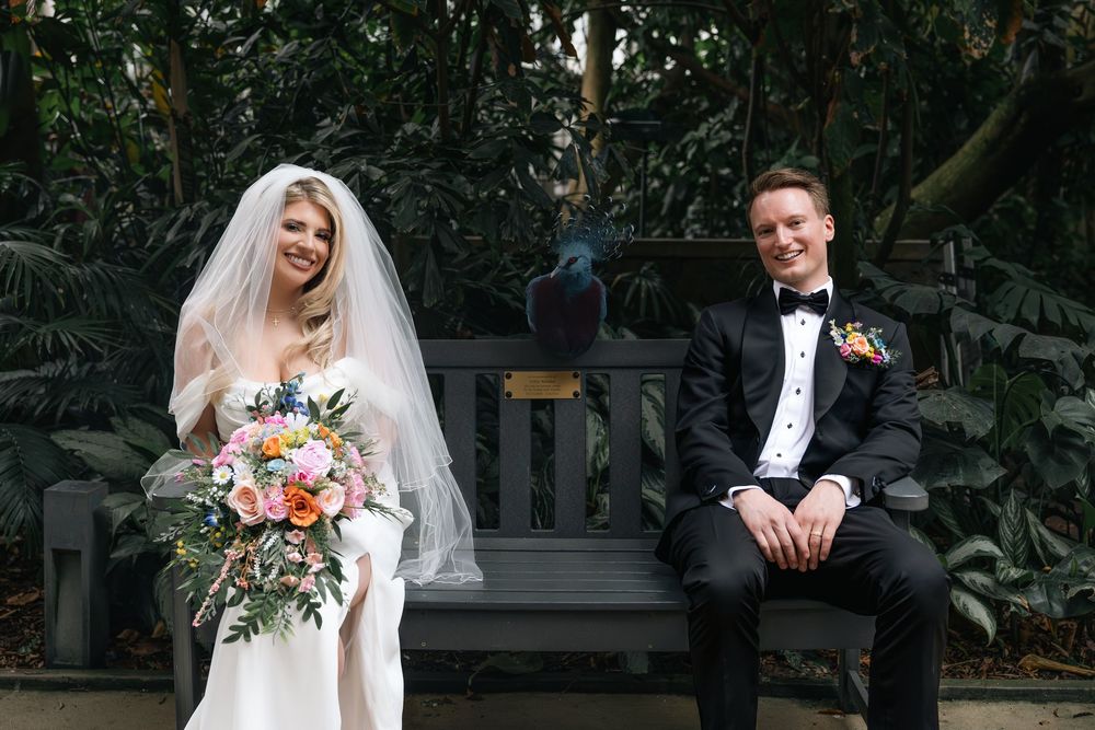 A bride and groom share a candid moment while sitting on a wooden bench surrounded by lush greenery.