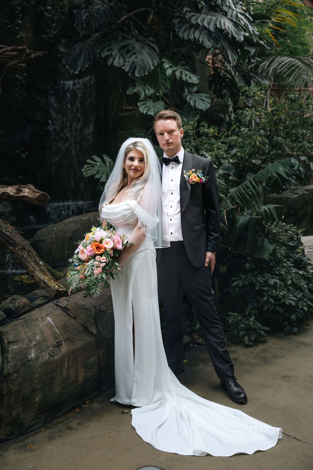 A wedding couple poses in front of lush greenery and tropical plants in an elegant botanical garden setting.