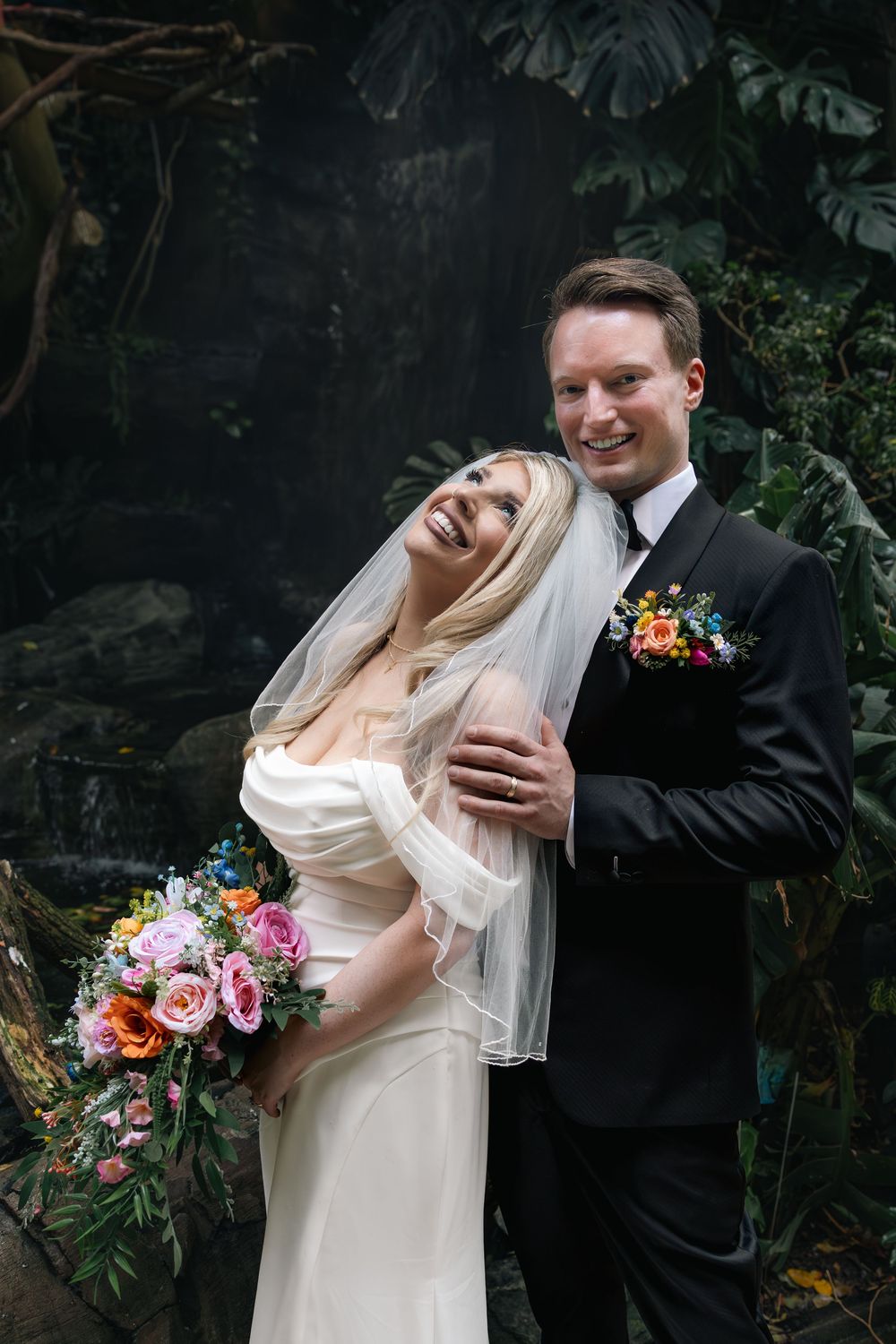 A joyful wedding couple shares a candid moment outdoors with a colorful bouquet against dark stone background.