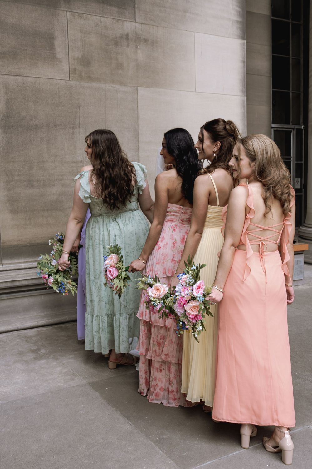 Row of bridesmaids in pastel dresses holding pink and white floral bouquets standing against a stone wall.
