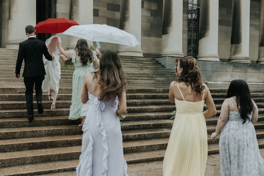 Wedding party climbs stone stairs in the rain with white and cream dresses as attendees hold umbrellas overhead.
