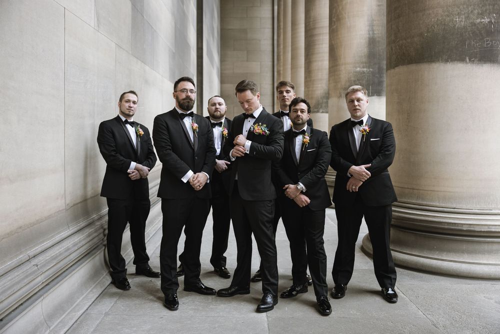 A group of groomsmen in black tuxedos pose together against white stone columns in an elegant setting.