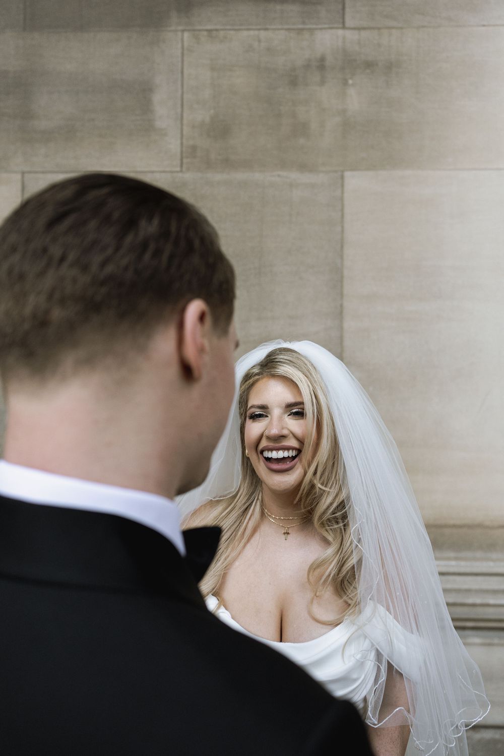 Bride wearing flowing white veil laughs joyfully while interacting with groom during wedding photoshoot.