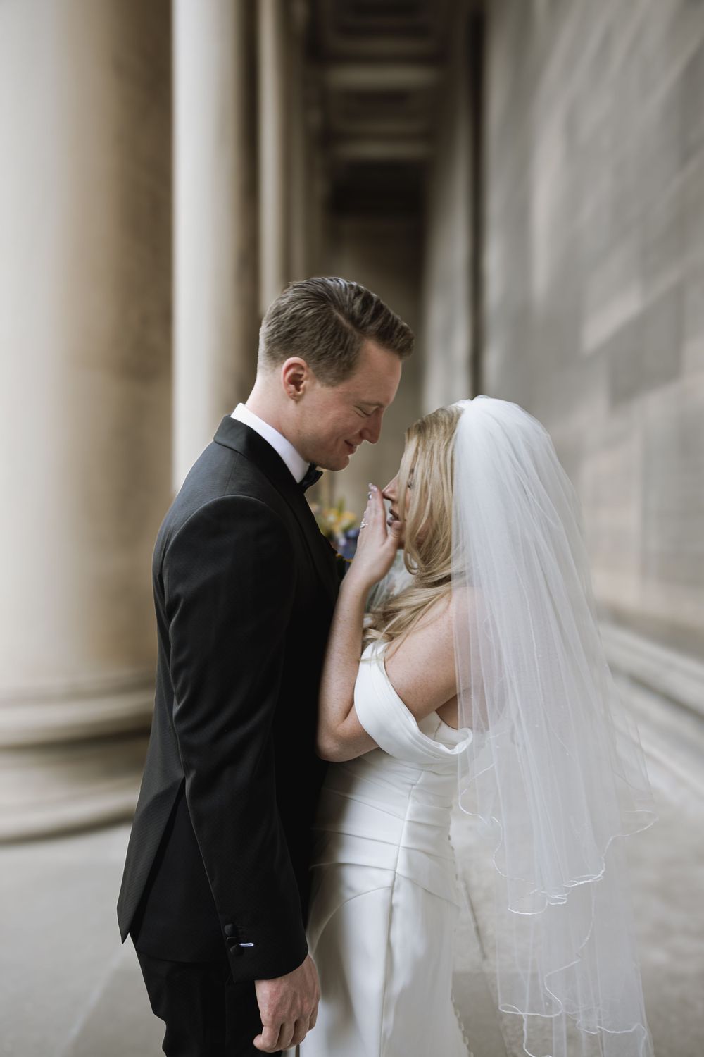 A newly married couple shares an intimate moment in a hallway with classical columns, the bride wearing a white gown and veil.