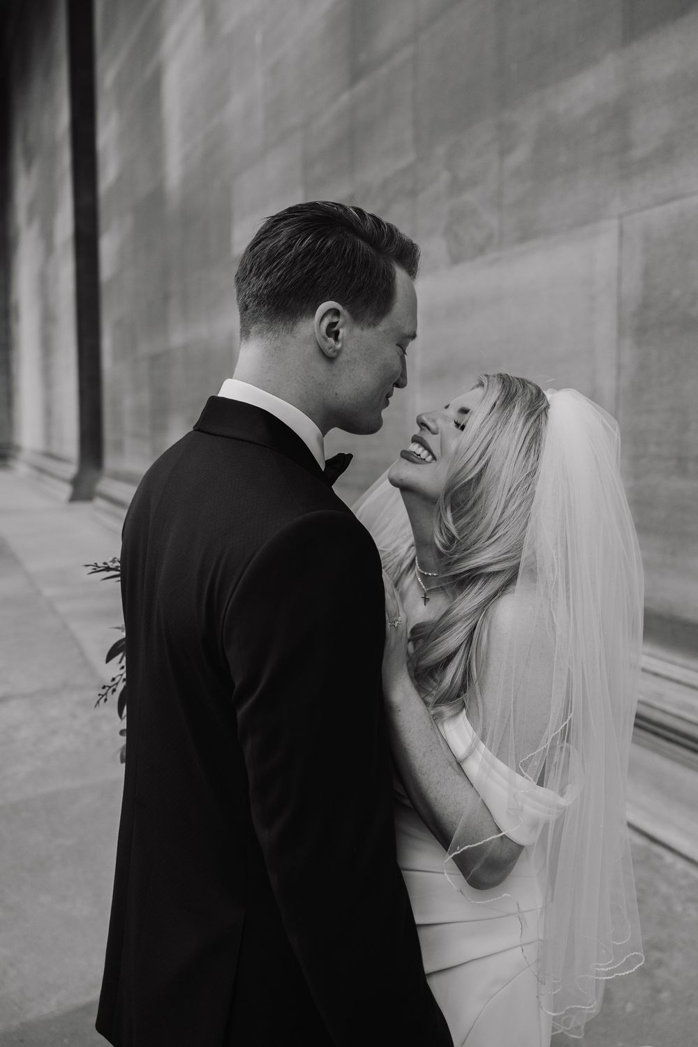 A romantic black and white wedding photo of a bride and groom sharing an intimate moment against a light wall.