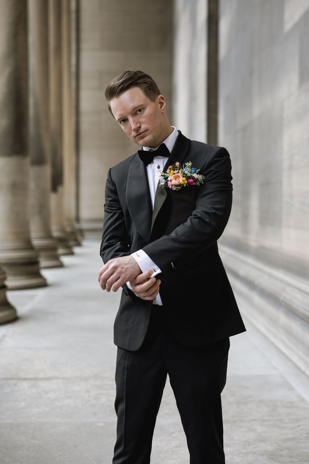A groom in a classic black tuxedo with boutonniere adjusts his sleeve against a backdrop of elegant stone columns.