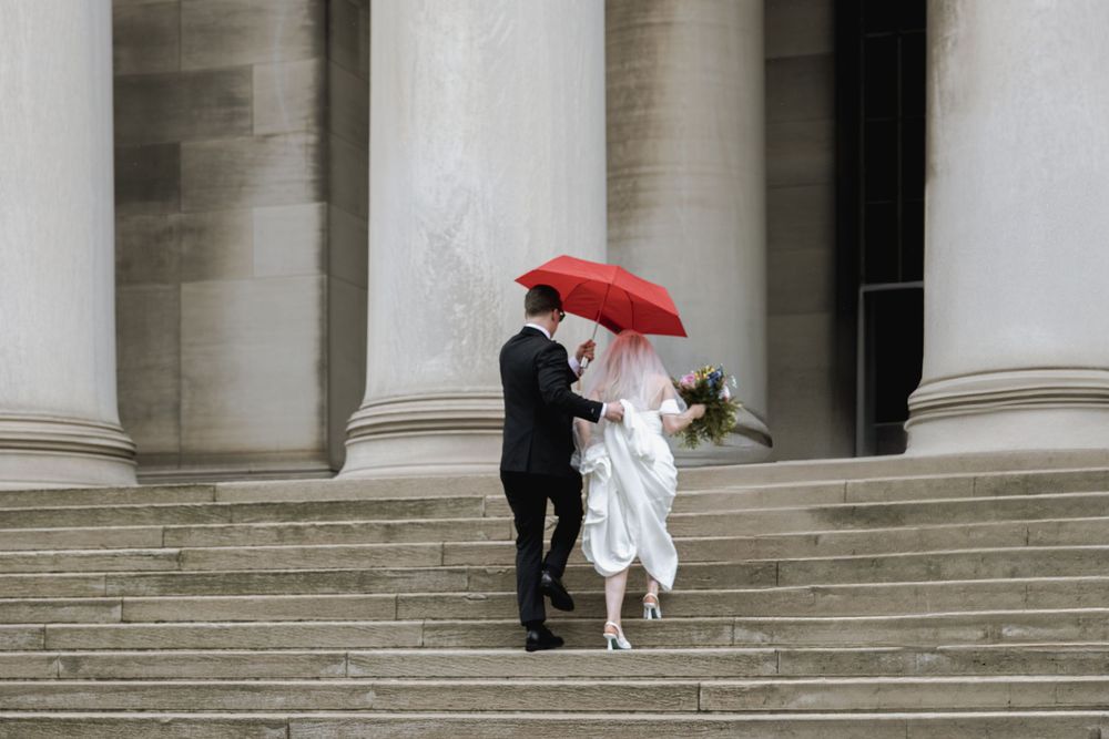 A couple stands on stone steps with a red umbrella against white pillared architecture in an elegant wedding setting.