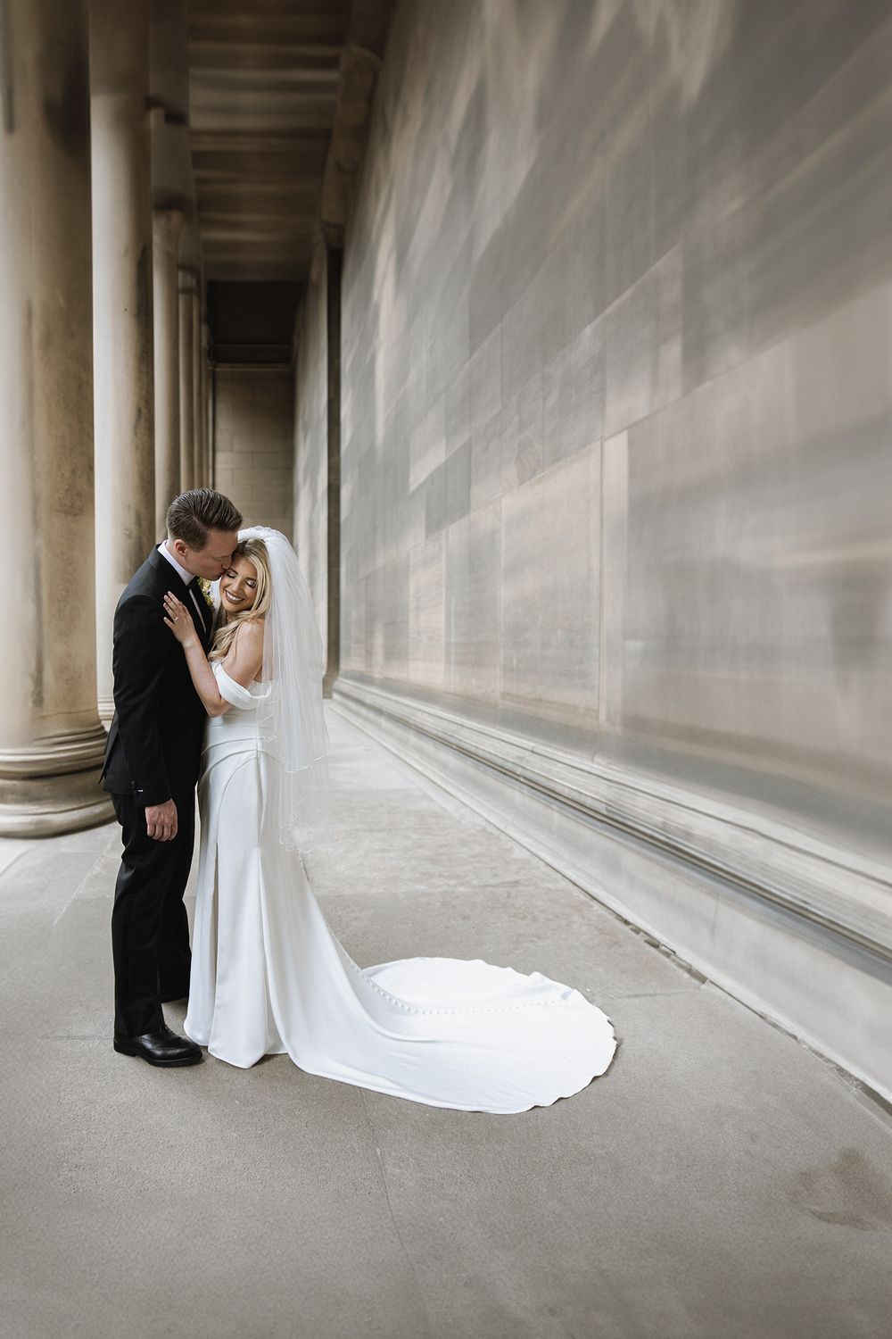 A couple shares an intimate moment in an elegant columned hallway, with the bride wearing a flowing white gown with train.
