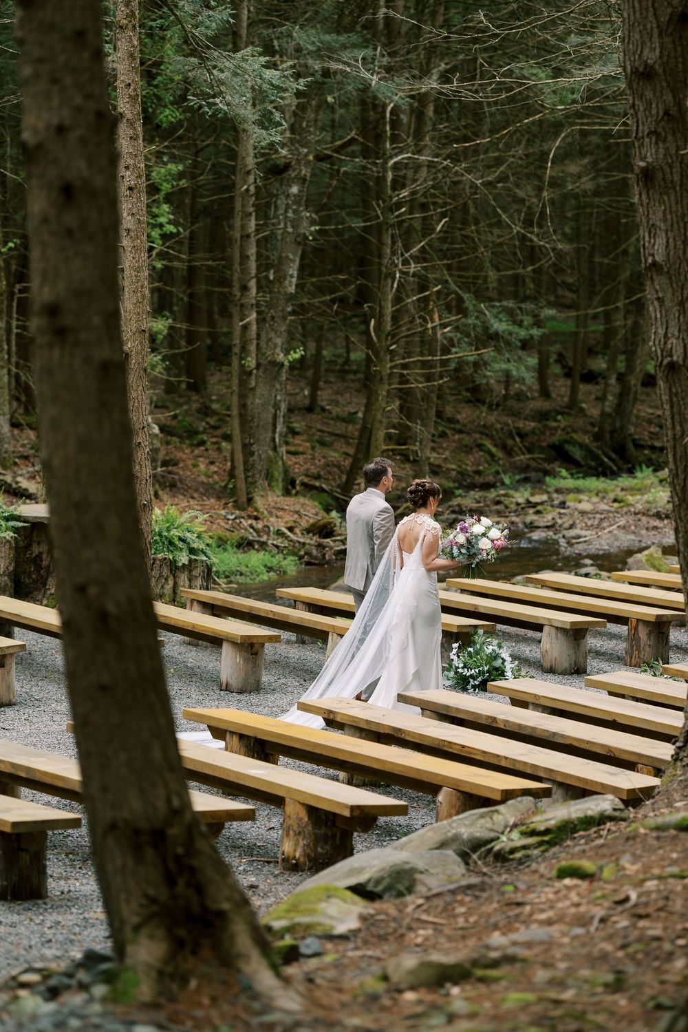 A couple in wedding attire stands on wooden benches in a peaceful forest setting surrounded by tall trees at Windham Manor.