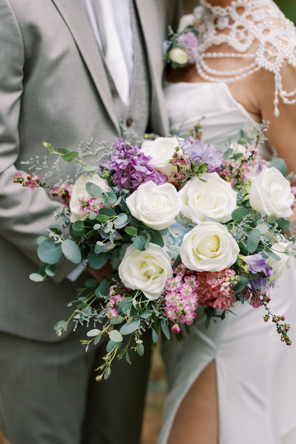 A wedding bouquet with white roses, purple lilacs, and eucalyptus greens held against a light gray suit at Windham Manor.