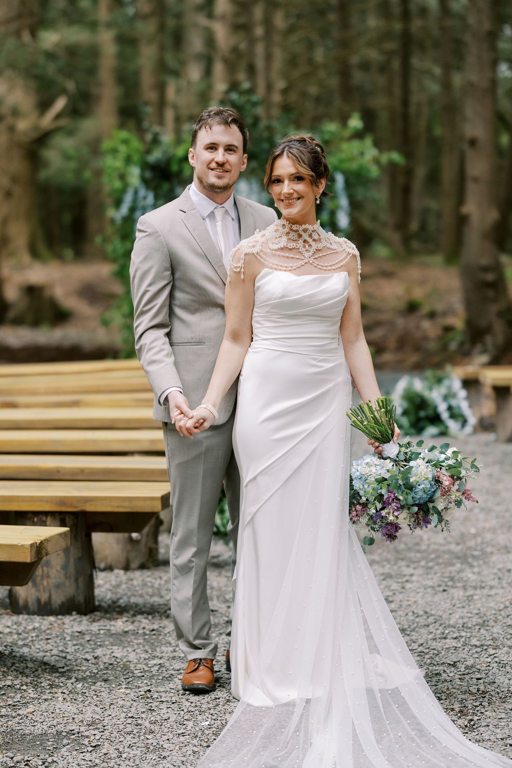A couple poses in the woods for their wedding photo, wearing a light gray suit and elegant white lace gown at Windham Manor.