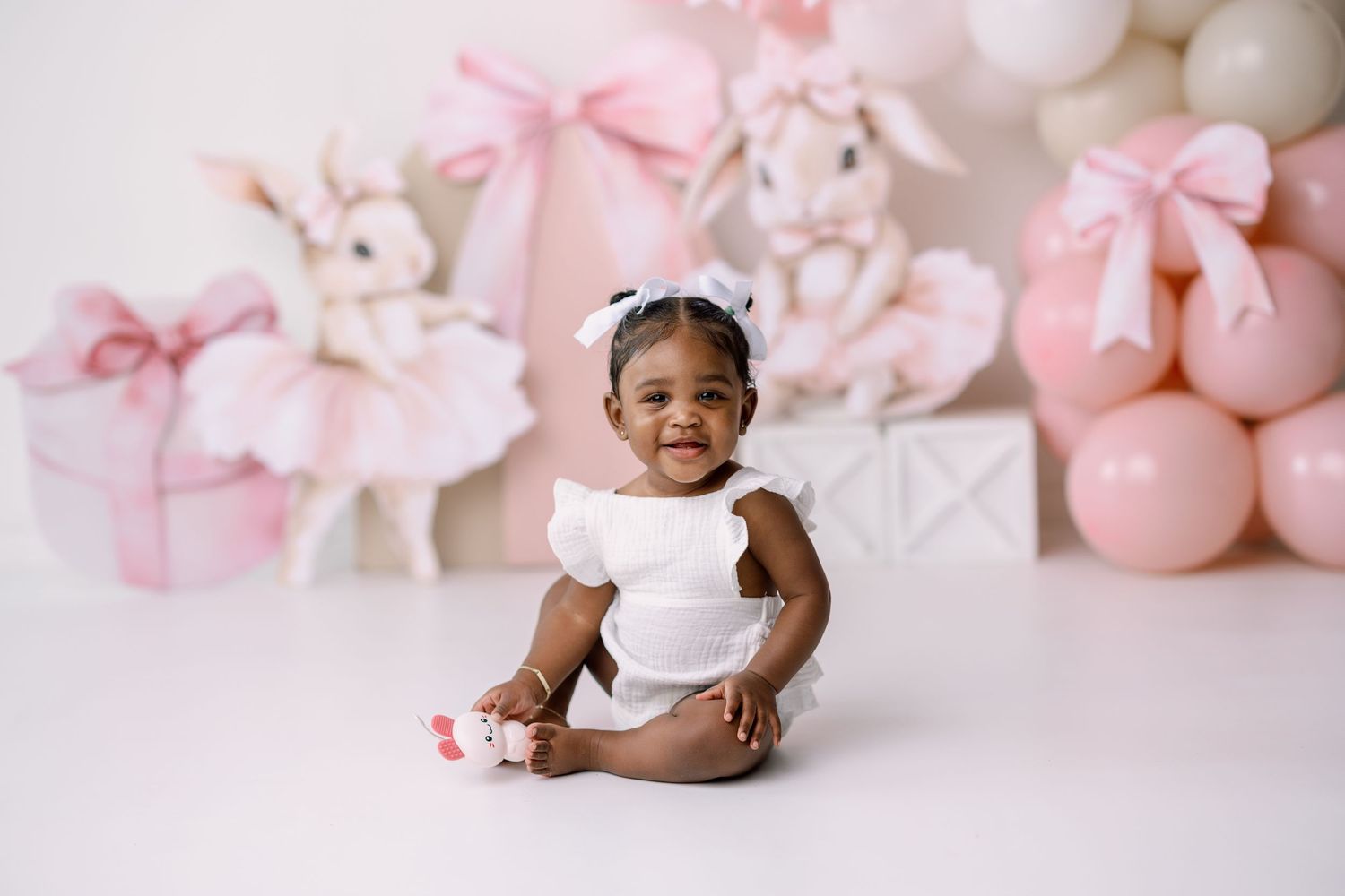Baby in white romper sits on pink backdrop surrounded by plush toys and pink bows for studio portrait session.