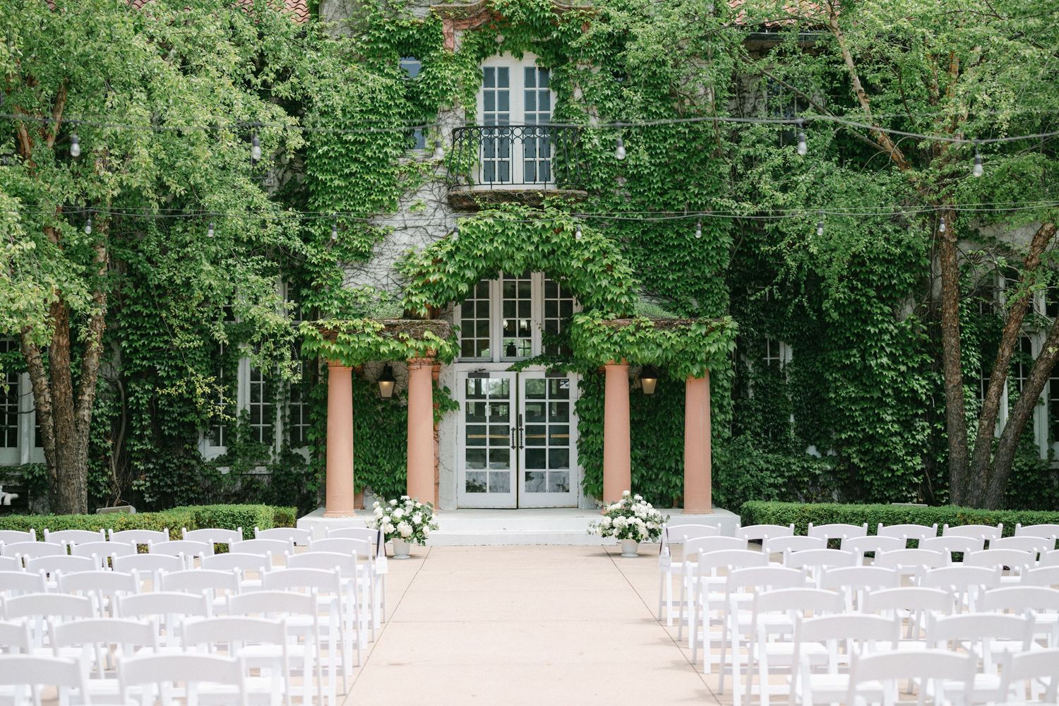 An elegant wedding ceremony setup with white chairs facing a vine-covered mansion with coral pillars and French doors.