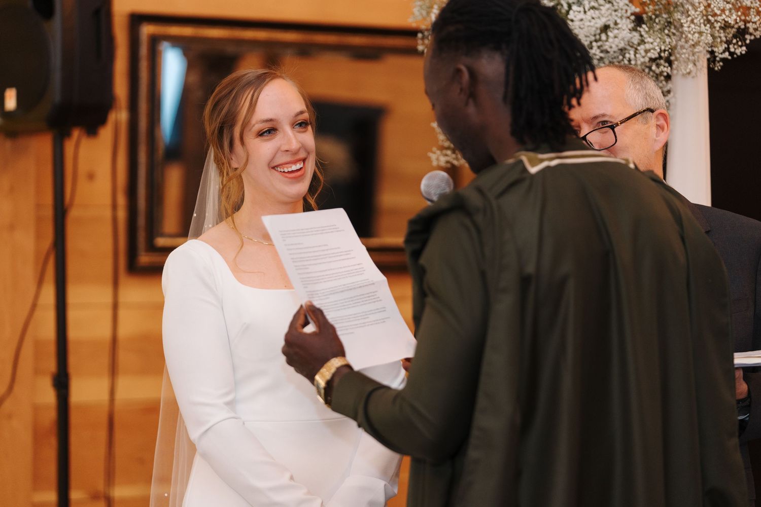 Smiling bride reading from paper during wedding ceremony under floral decorations.