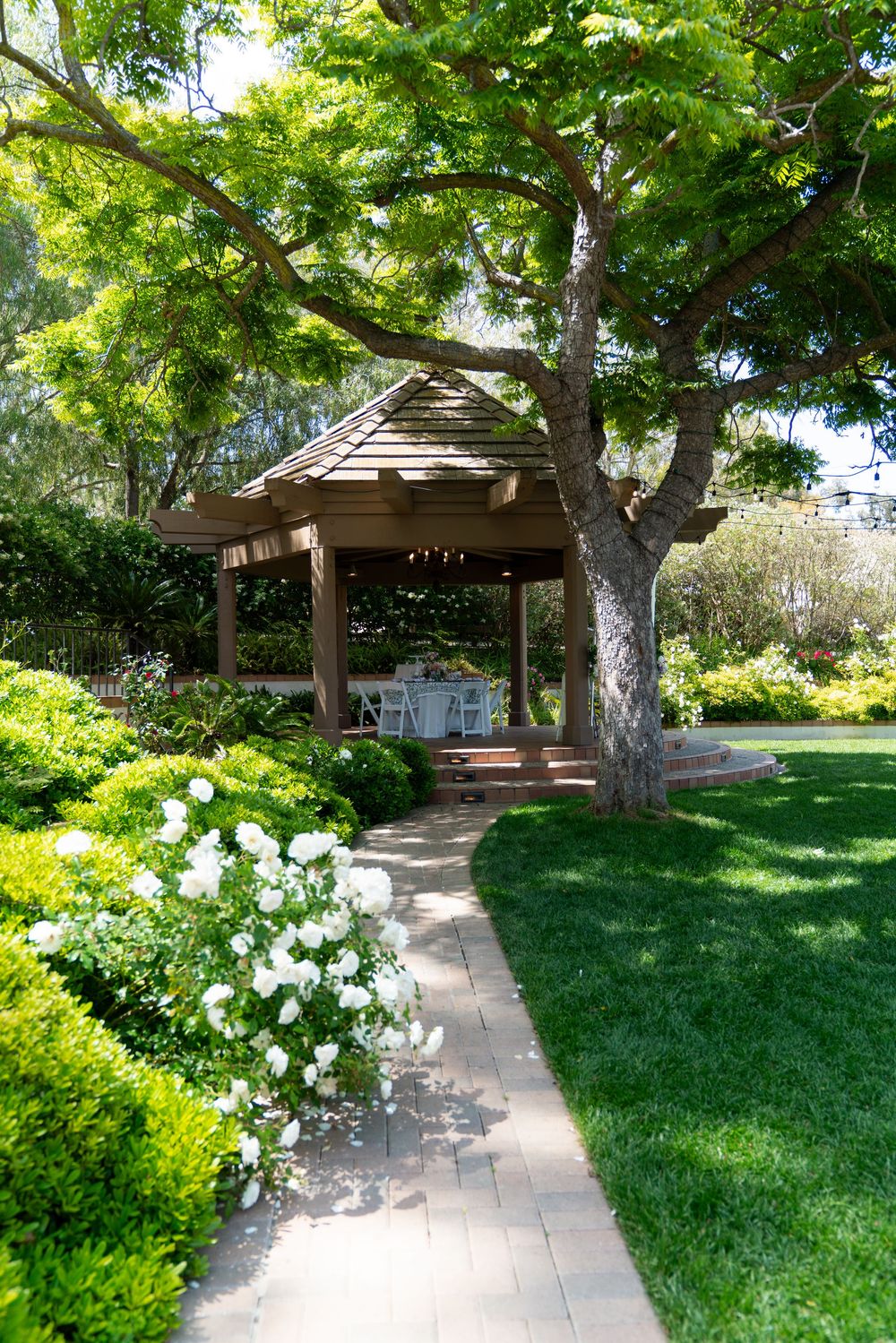 A wooden gazebo surrounded by lush gardens and trees with a stone pathway leading to it.