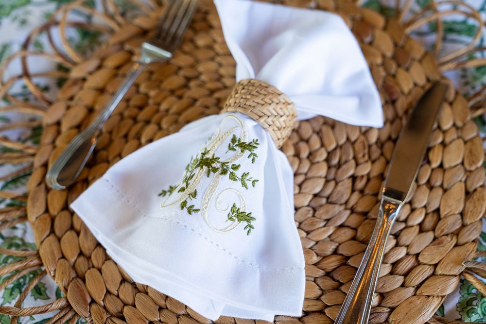 White linen napkin with green embroidered herbs placed on a woven rattan placemat with silverware.