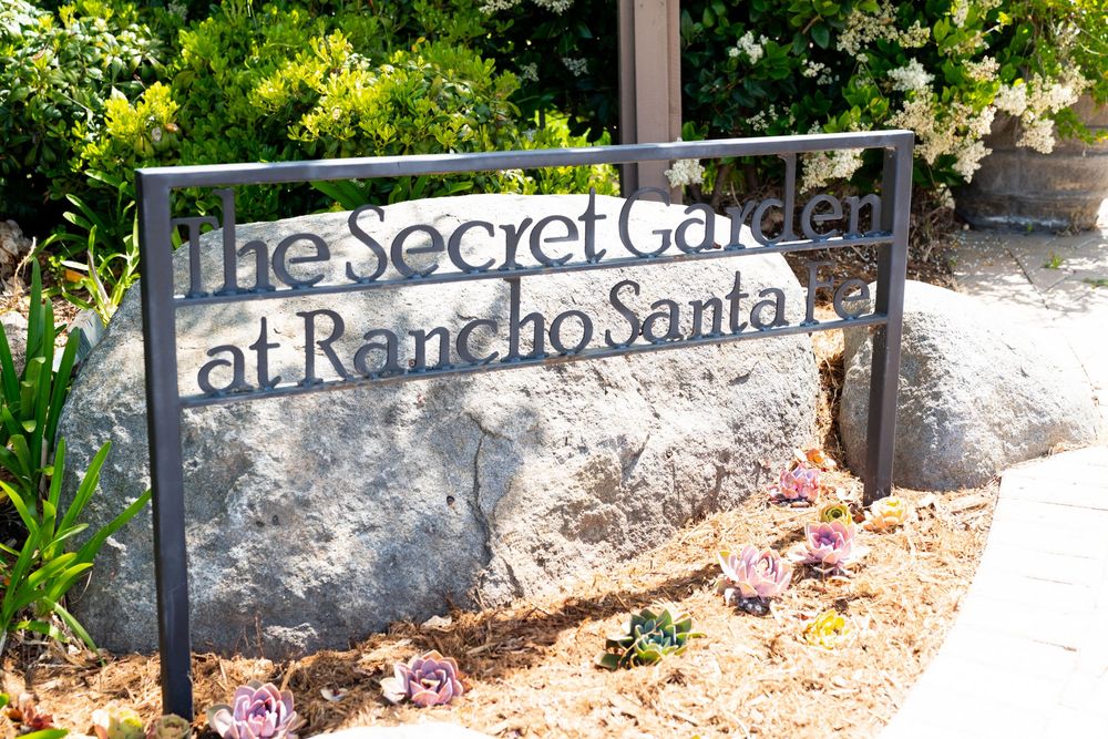Metal sign reading 'The Secret Garden at Rancho Santa Fe' mounted on a stone wall with greenery in the background.