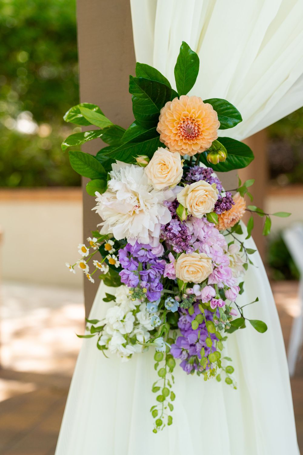 Beautiful floral arrangement with peach dahlias, white peonies, cream roses and purple delphinium on white drapery.