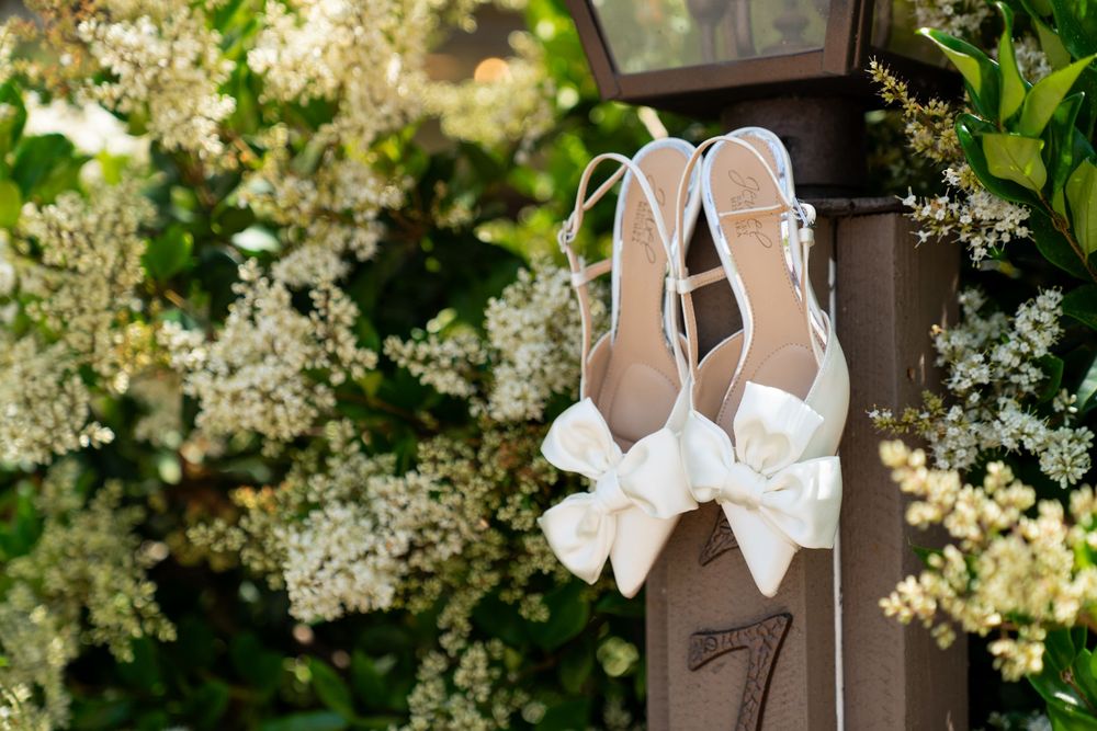 White wedding shoes with bows hanging on wooden post surrounded by delicate white flowers and greenery.